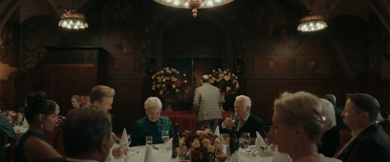 Peacock (2010) movie still: wide — A dark wood-paneled hall features high ceilings and ornate wall murals. Guests sit at round dinner tables…