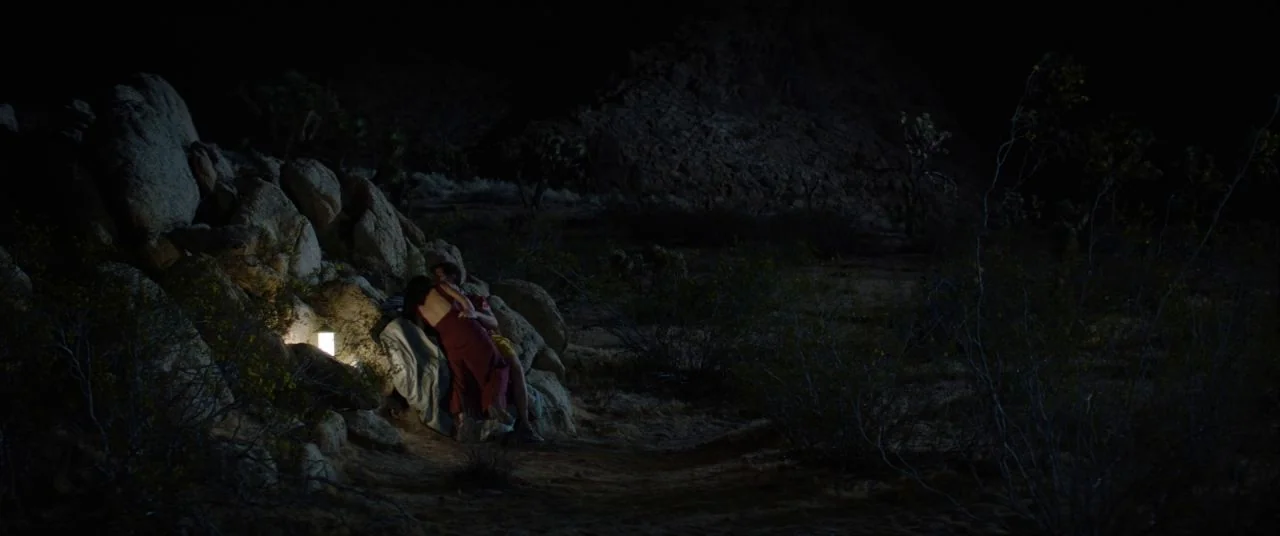 Palm Springs (2020) movie still: wide — A dark, rocky desert landscape stretches out under a night sky. A man and woman sit huddled together against…