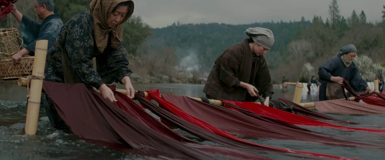 Memoirs of a Geisha (2005) movie still: wide — A group of people work along a shallow riverbank under a muted cloudy sky. They rinse and manipulate…