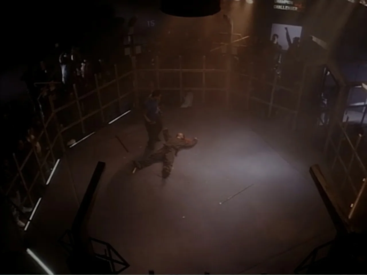 Firepower (1979) movie still: wide — A dimly lit industrial fighting enclosure with a wooden framework perimeter and concrete floor. One man stands…
