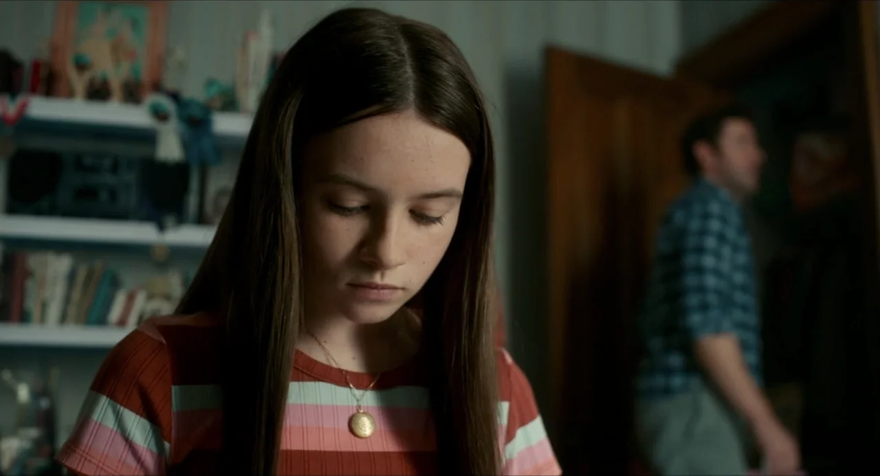 IF (2024) movie still: closeup — A bedroom features a cluttered bookshelf behind a young girl with long brown hair. The girl looks downward while an…