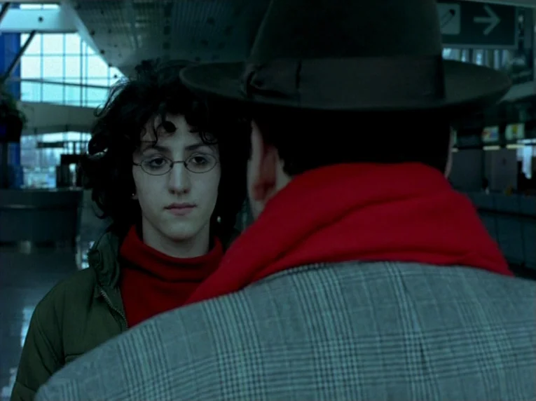 Notre Musique (2004) movie still: medium — A spacious airport terminal with large glass windows and an overhead directional sign. A young woman…