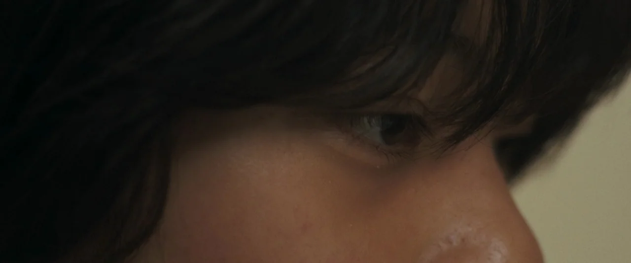 Monster (2023) movie still: closeup — A close-up view shows a person's eye and temple area in soft light. The dark hair frames the upper portion of…