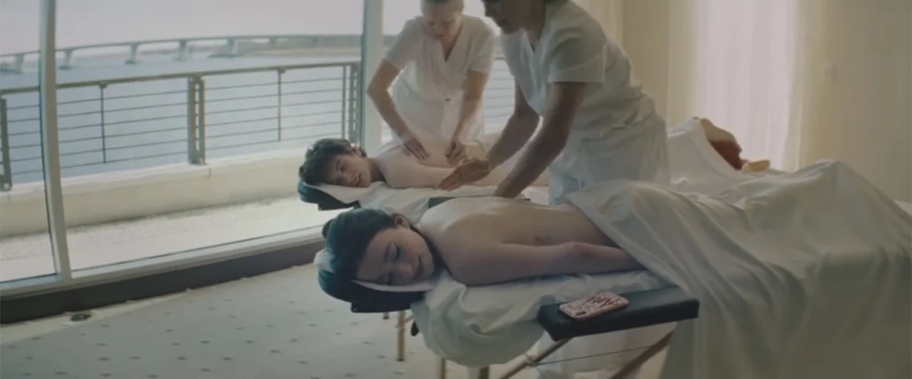 Anora (2024) movie still: medium — A bright spa room features two massage tables arranged side by side near a window. Two therapists in white…