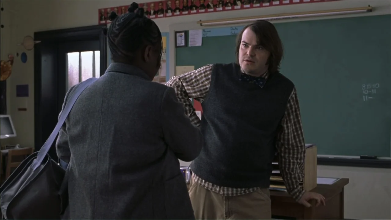 School of Rock (2003) movie still: medium — A school room contains a chalkboard covered in papers and a wooden desk. A man wearing a sweater vest…