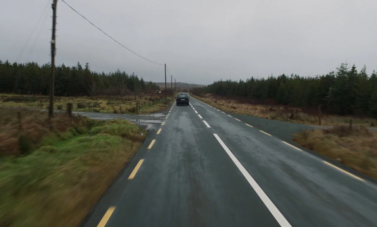 It Is In Us All (2022) movie still: wide — A rural asphalt road cuts through a landscape of pine trees and moorland. A dark hatchback vehicle moves…