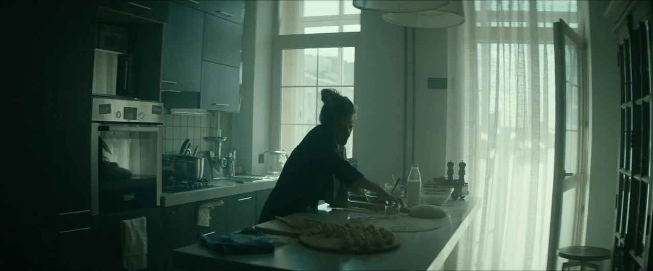 Invasion (2020) movie still: medium — A modern, minimalist kitchen features dark cabinetry and a central island with prepared dough. A woman stands…