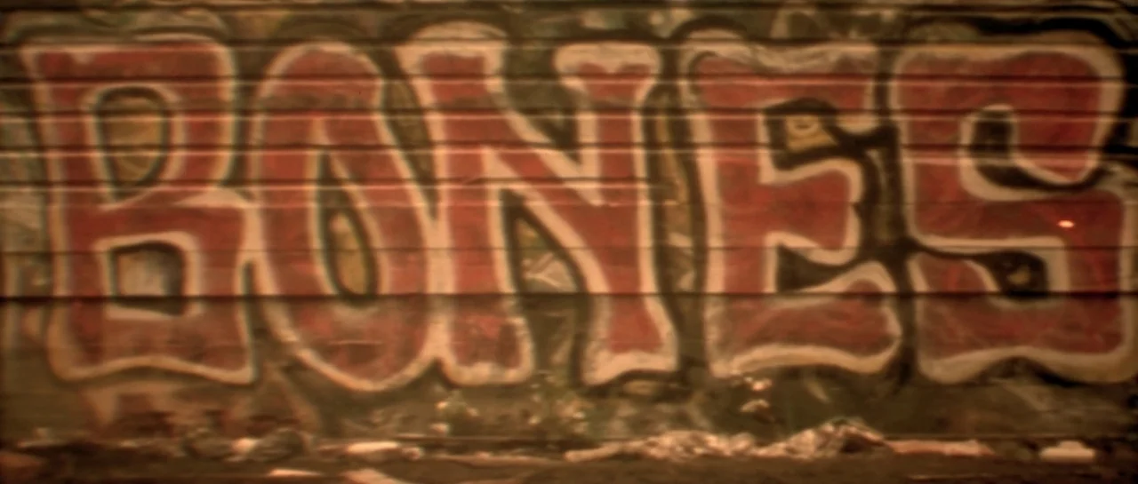 Bones movie still: closeup — A metal corrugated wall features large, faded graffiti lettering spelling out the word BONES. Trash and debris are…