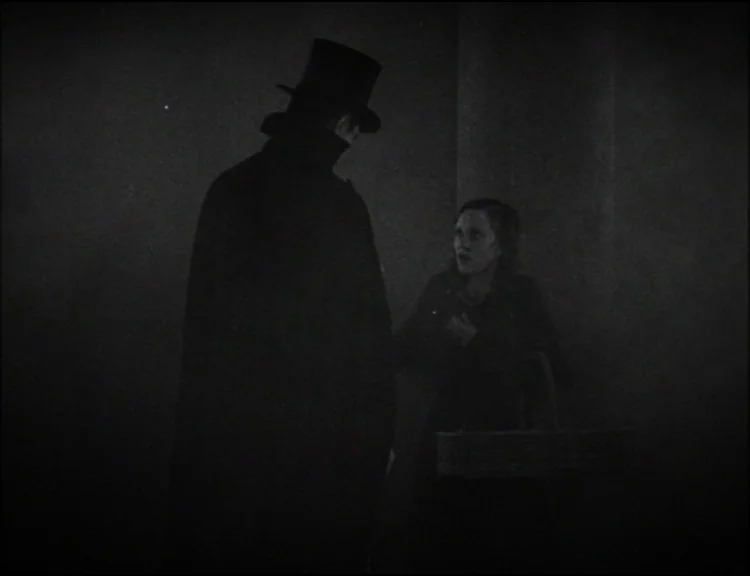Dracula (1931) movie still: medium — A dark, misty alleyway at night with deep shadows surrounding two figures. A man in a top hat confronts a woman…