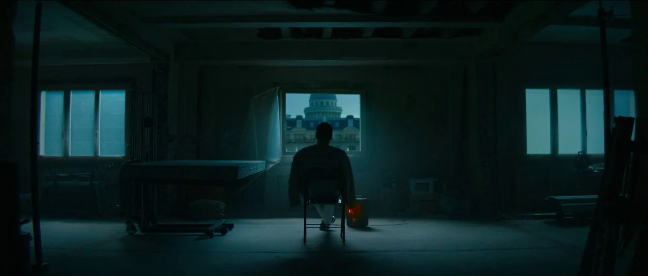 The Killer (2023) movie still: wide — A dimly lit room with unadorned walls and a low-slung ceiling structure. A person sits on a folding chair…