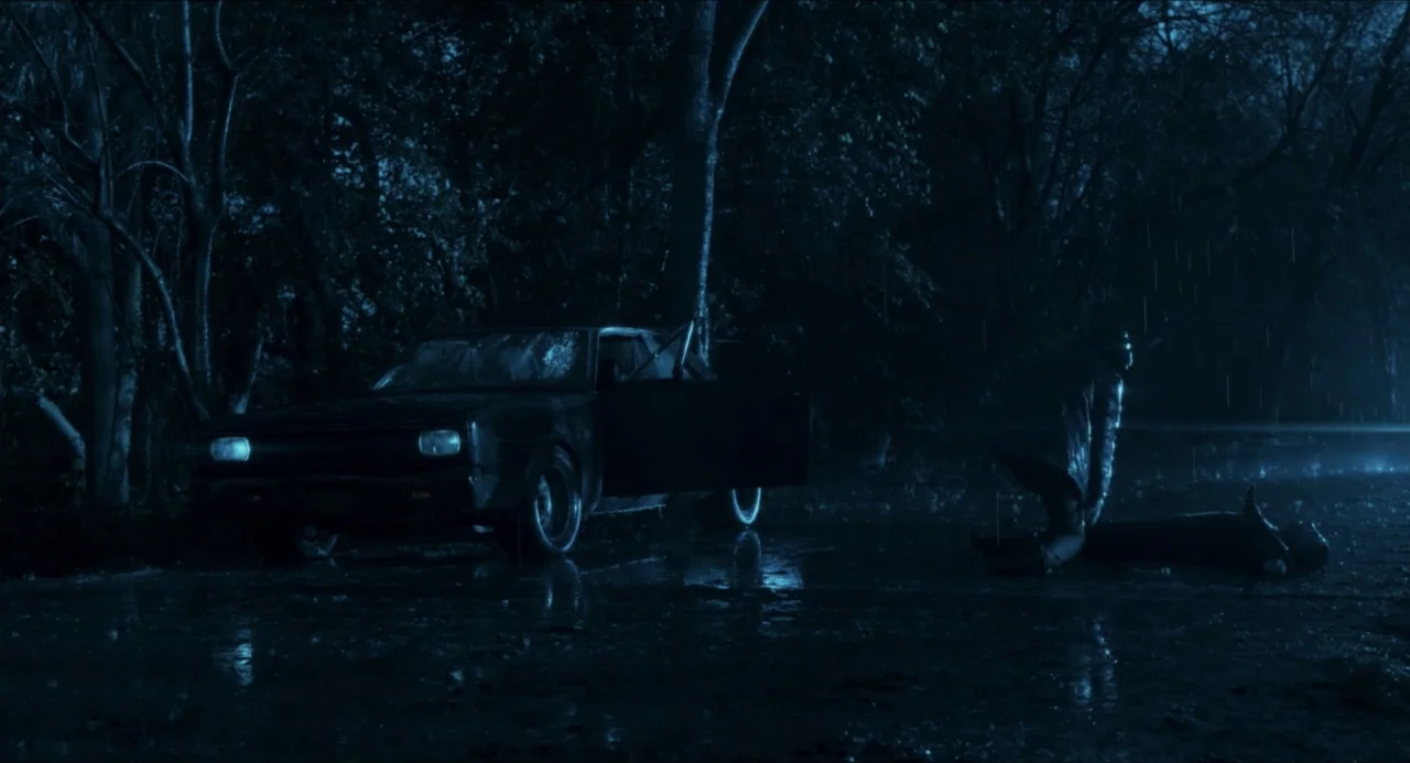 Echoes Of A Crime (2025) movie still: wide — A dark, rain-slicked road runs through a forest at night with a parked car. One person kneels beside…