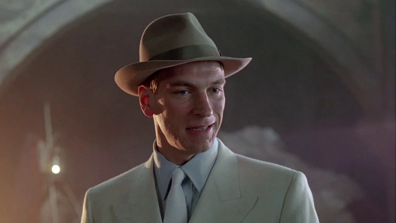 Naked Lunch (1991) movie still: medium — A dimly lit interior space with arched architectural elements visible in the background. A man in a light…