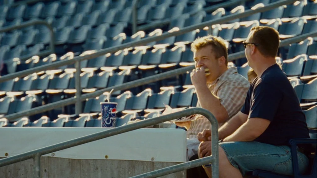 Sugar (2008) movie still: medium — A nearly empty sports stadium features rows of dark blue plastic seats under sunlight. Two men sit in the lower…