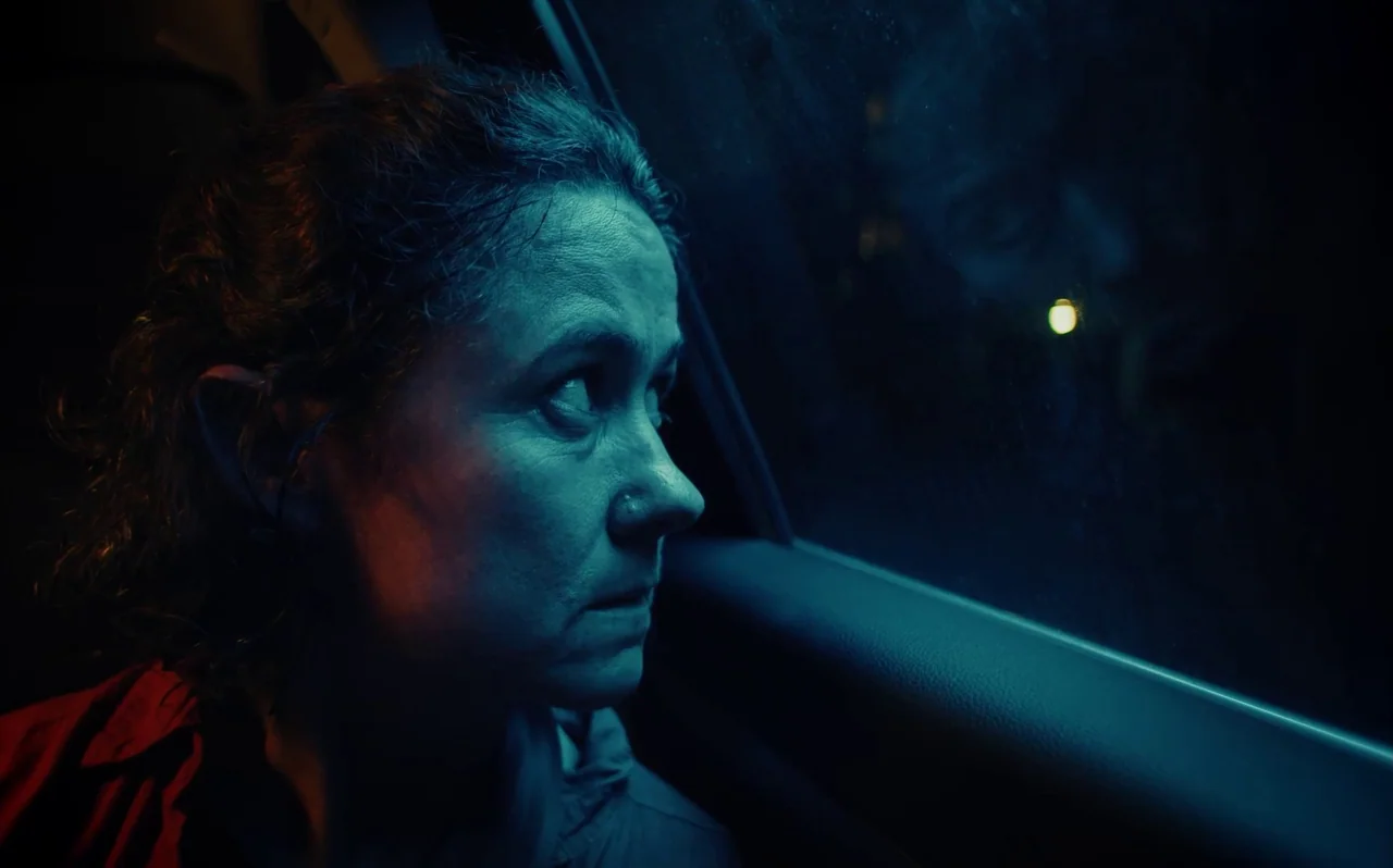 Knocking (2021) movie still: closeup — Inside a dark car cabin at night with a window view of blurred urban lights. A woman stares intently out of…
