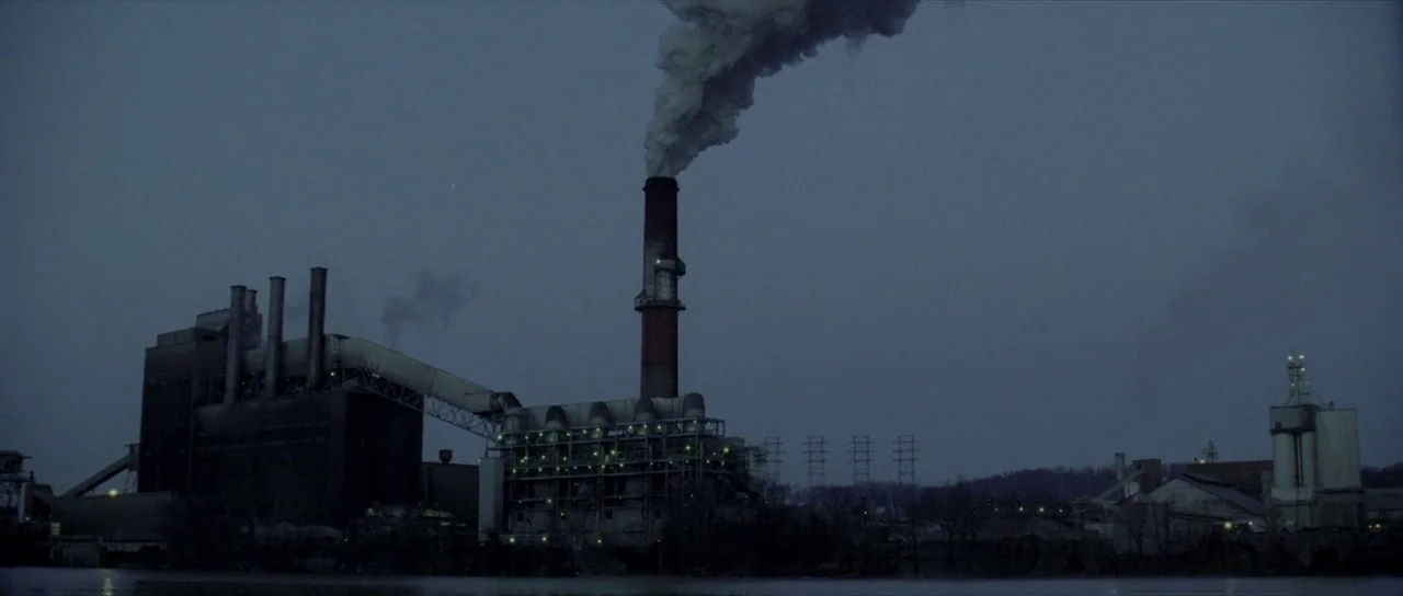 The Mothman Prophecies (2002) movie still: establishing — An industrial manufacturing facility sits near a dark body of water at dusk. A tall…