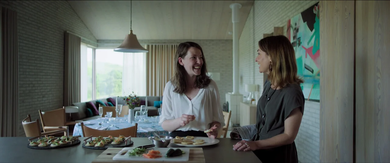 The Feast (2021) movie still: medium — A modern kitchen with light-colored brick walls and large windows looking toward an open living area. Two…