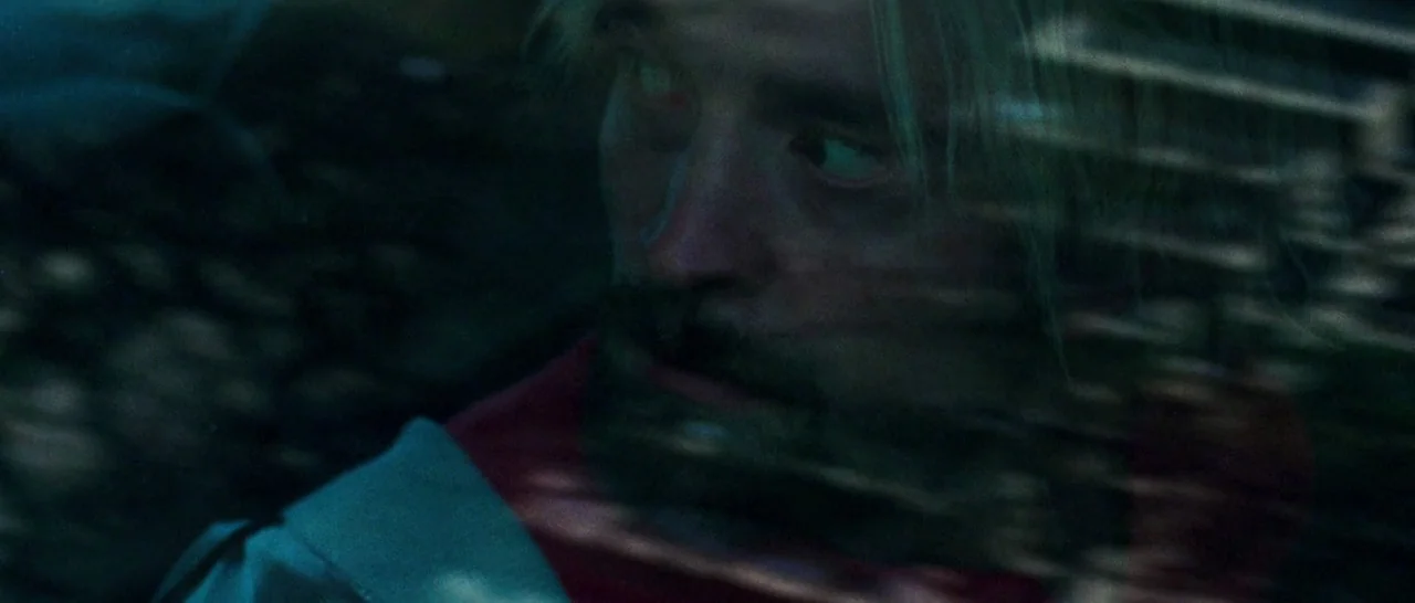 Good Time (2017) movie still: closeup — A man sits inside a vehicle with reflections visible on the dark glass surface. The subject peers through…