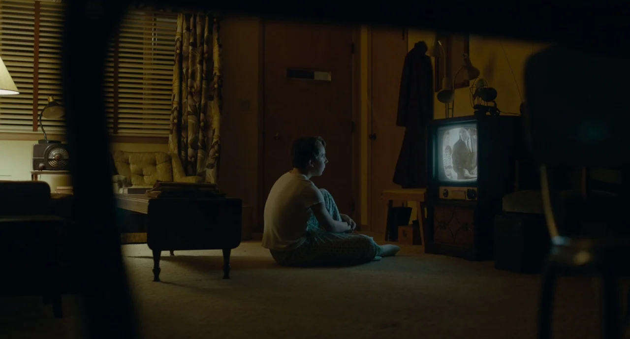 Wildlife (2018) movie still: wide — A dark living room features a patterned curtain, a vintage radio, and carpeting. A young boy sits on the floor…