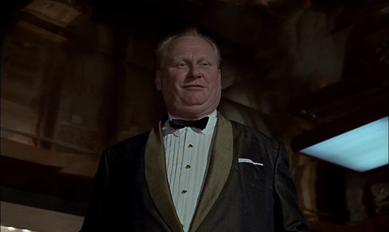 Goldfinger (1964) movie still: medium — An older man wearing formal evening attire stands in a dimly lit, dark space. The subject stares directly…