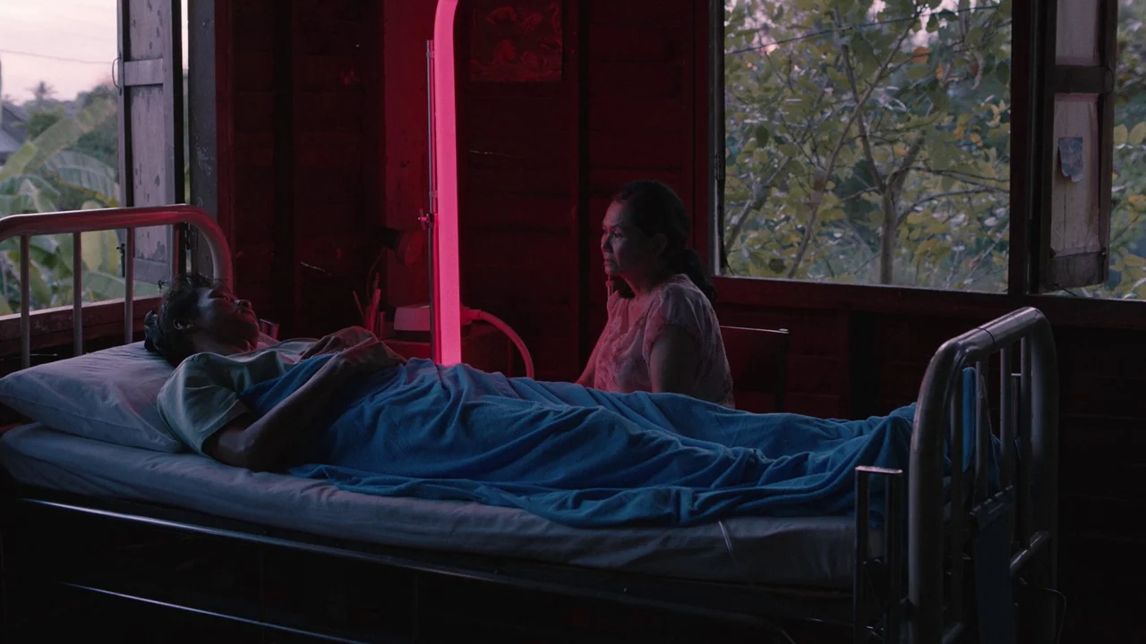 Cemetery of Splendour (2015) movie still: medium — A dimly lit room with wood-paneled walls features a single metal hospital bed. A woman sits on a…
