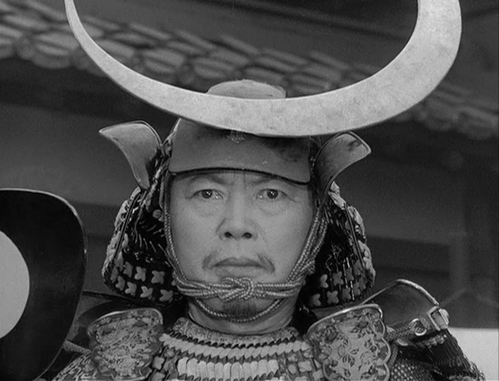 Throne of Blood (1957) movie still: closeup — An outdoor area features traditional Japanese roof tiles visible in the blurred background. A man…