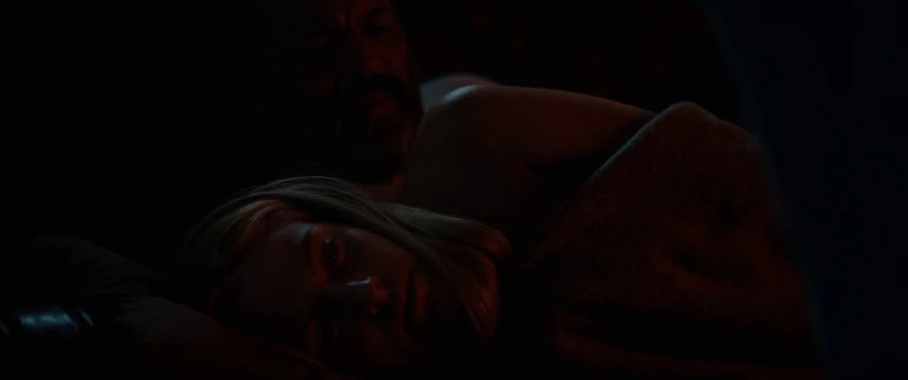 Hold The Dark (2018) movie still: closeup — A dark, dimly lit indoor setting where two people lie in bed. A man and a woman are huddled together…