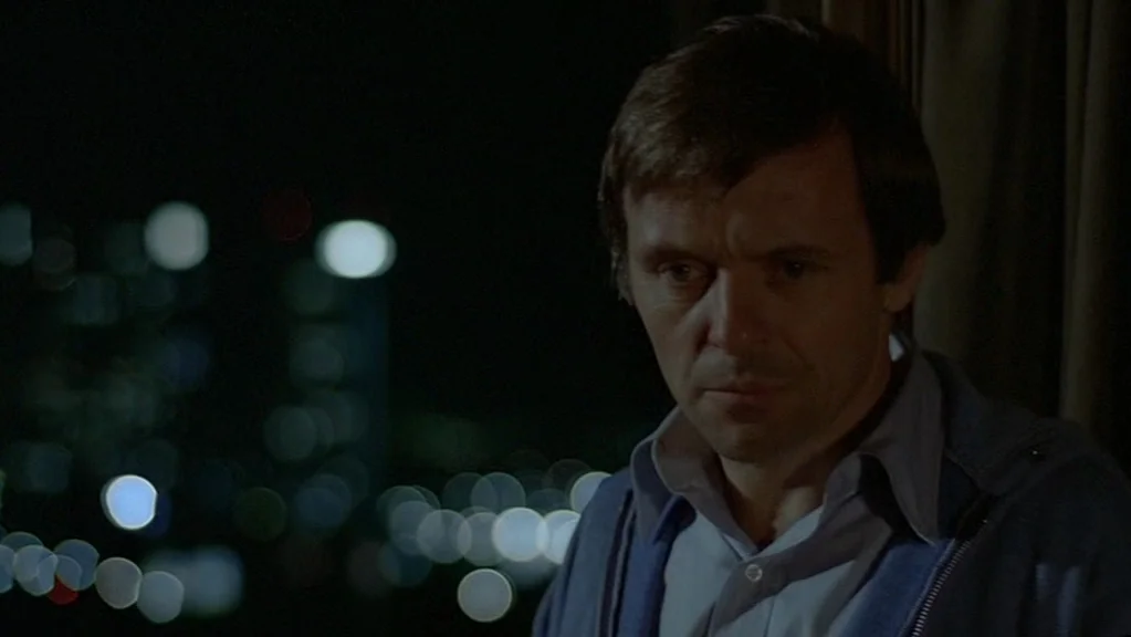 Magic (1978) movie still: closeup — A man stands indoors at night looking toward a window with blurred city lights. The dark room interior is…