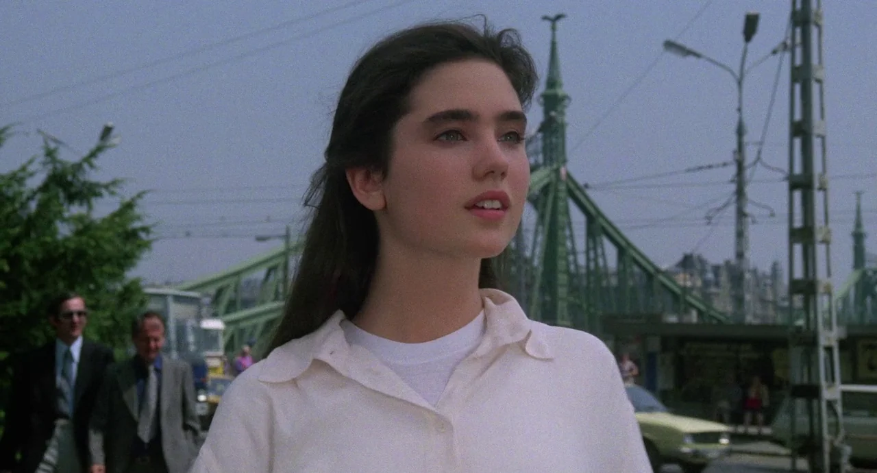 Etoile (1989) movie still: medium — A woman stands in an urban setting with a large green metal bridge behind her. Two men in business suits walk in…