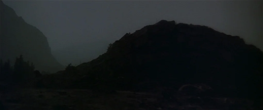 Clan of the Cave Bear (1986) movie still: establishing — A dark rocky mountain range stands against a hazy night sky. Sparse pine trees grow at the…