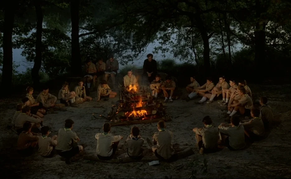 Murmur of the Heart (1971) movie still: wide — A group of boys wearing scouting uniforms gather around a fire in a dense woods. The scouts sit on…