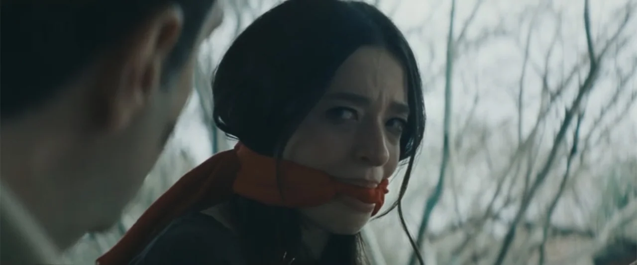 Anora (2024) movie still: closeup — A dense forest with leafless trees creates a cold and muted background environment. A young woman with a red…