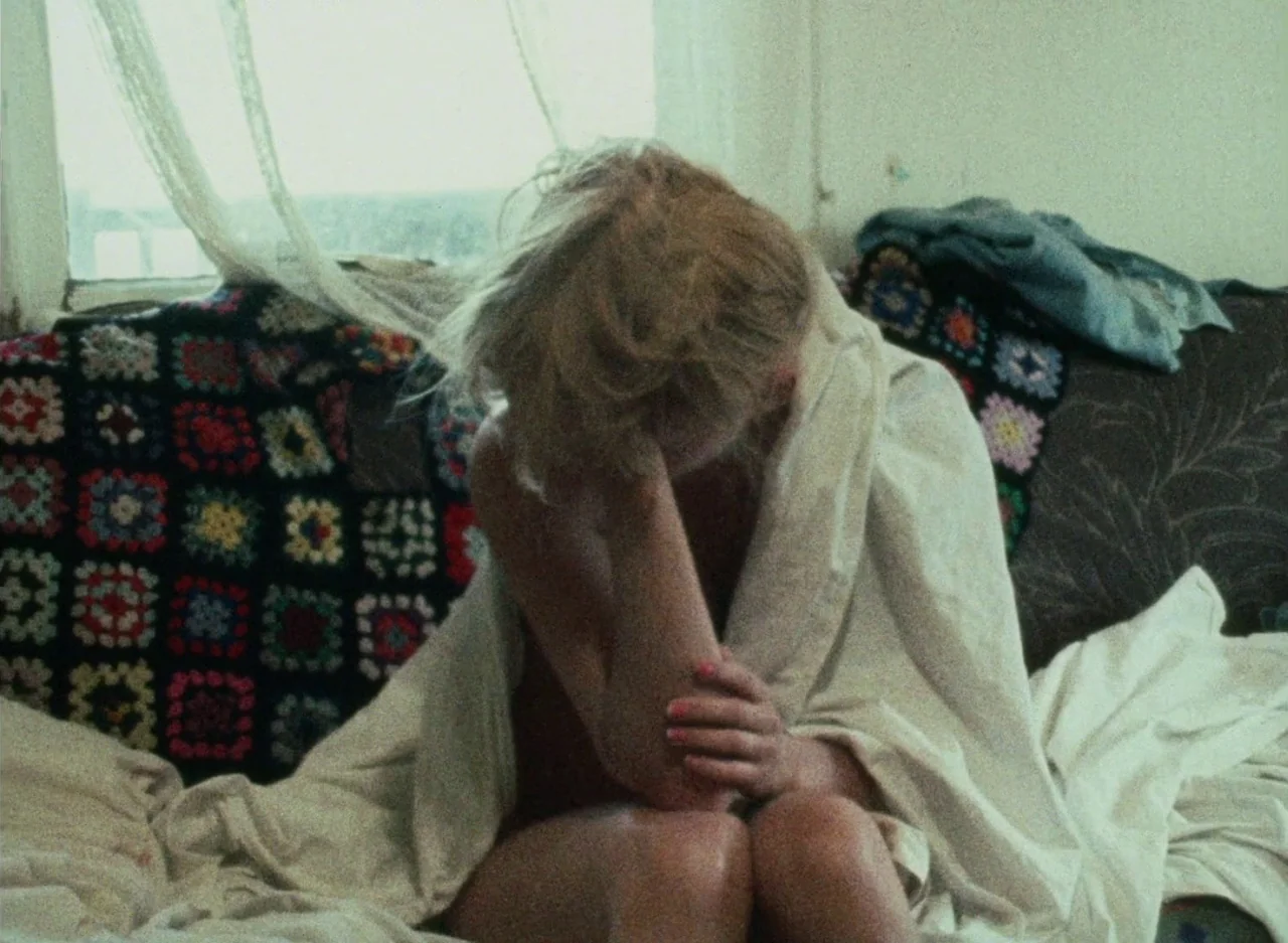 Wanda (1970) movie still: medium — A person sits on a sofa draped in a blanket near a window. A colorful crochet throw pillow sits beside them.
