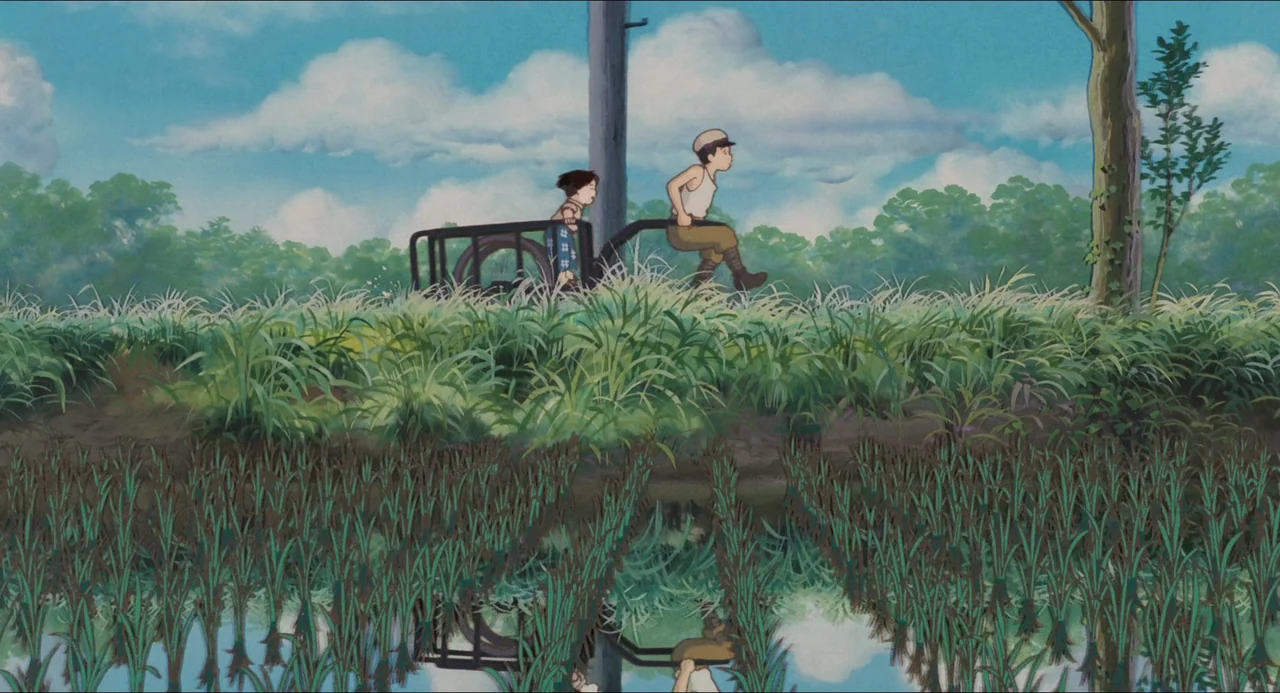 Grave of the Fireflies (1988) movie still: wide — A lush green rice paddy borders a path under a blue sky with clouds. A young boy pulls a small…
