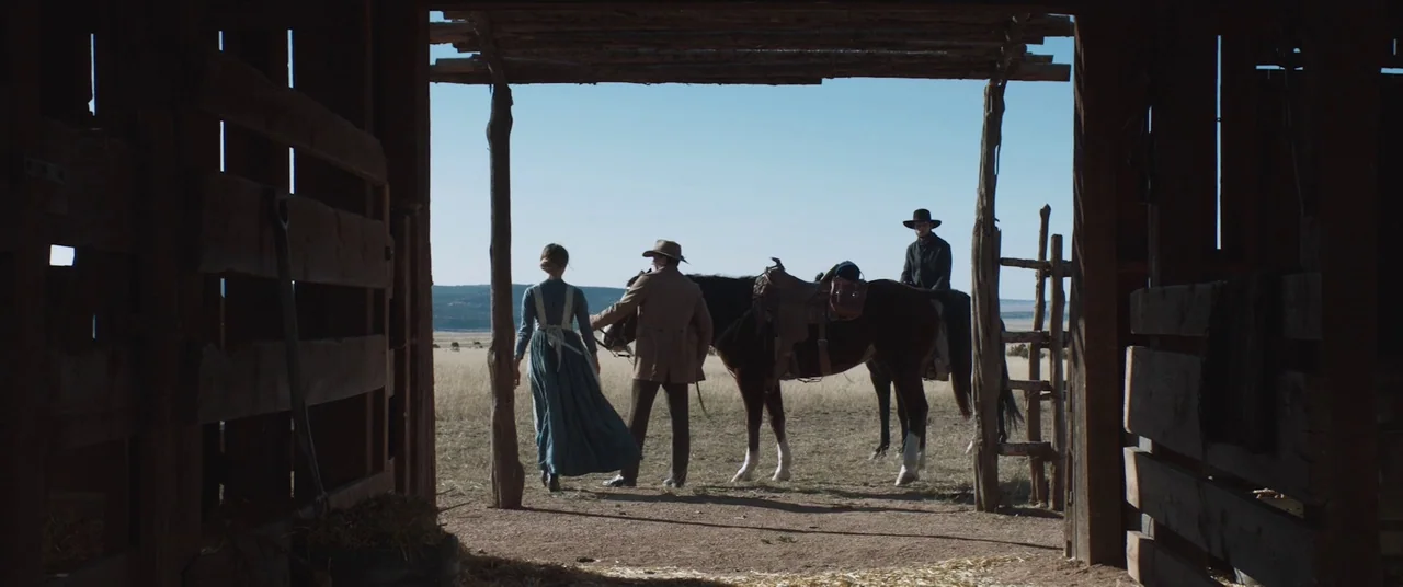 The Wind (2018) movie still: wide — A dark wooden barn interior frames a bright view of an expansive yellow field. A woman and two men stand near a…
