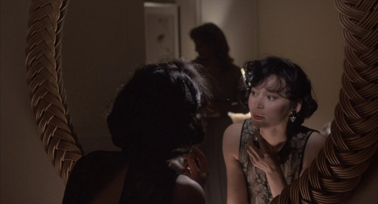 Masquerade (2023) movie still: medium — A dimly lit room contains a large circular mirror with a woven wicker frame. A young woman sits before the…