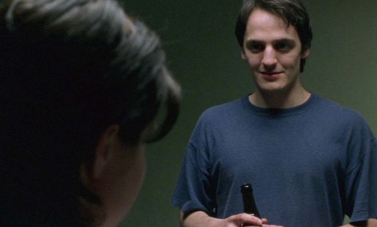 Rosetta (1999) movie still: medium — A dimly lit room with a muted green wall serves as the backdrop. A man holding a bottle looks toward a person…