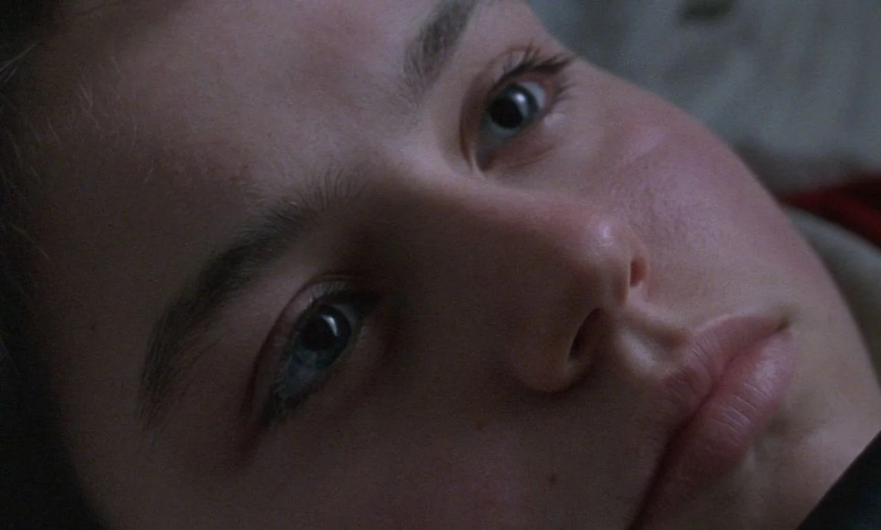 Rosetta (1999) movie still: closeup — A dark, dimly lit indoor environment with soft shadows across the subject's features. A young woman lies…