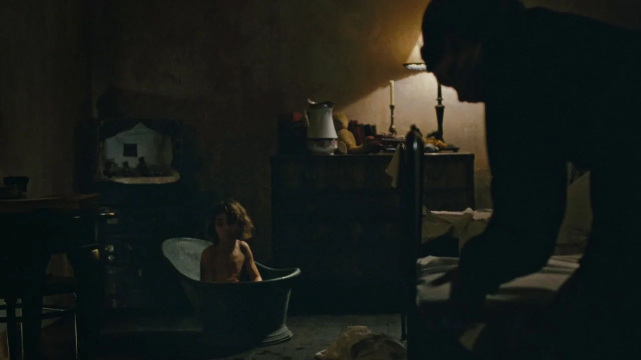 Childhood of a Leader (2015) movie still: medium — A dimly lit room contains an antique wooden dresser, a small tub, and a bed. A child sits in the…
