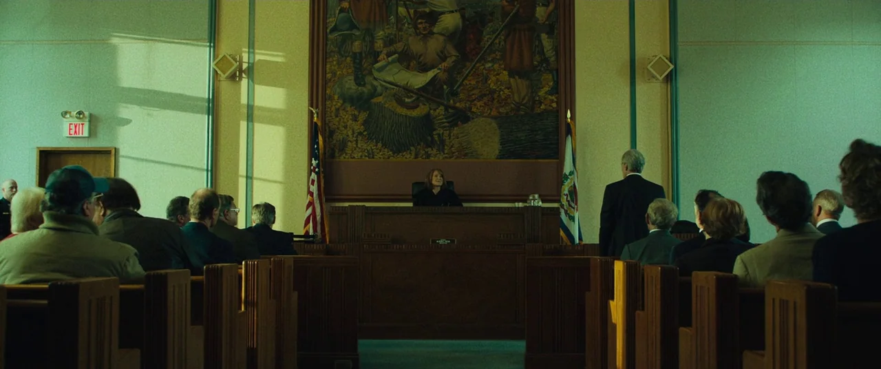 Dark Waters (2019) movie still: wide — A courtroom interior with high ceilings and rows of wooden pews facing a judge. A large historical mural is…