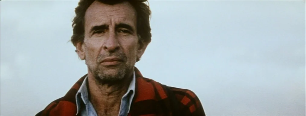 Japón (2002) movie still: closeup — A man stands outdoors under an overcast, featureless sky. He looks forward while wearing a dark red and black…