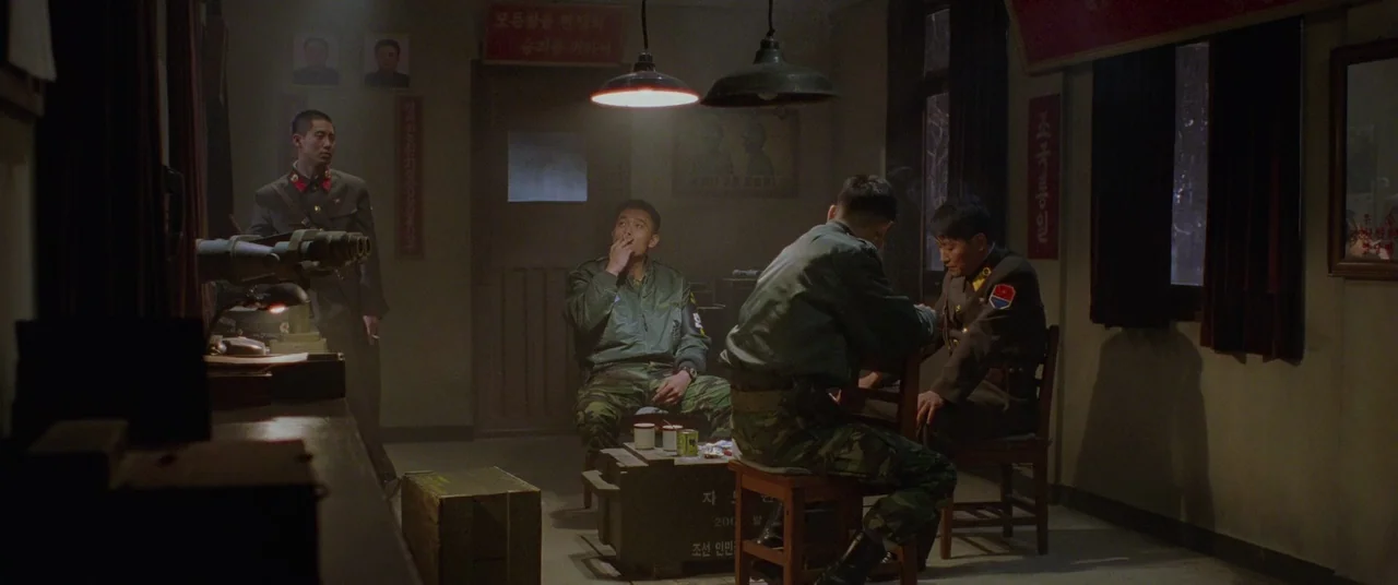 Joint Security Area (2000) movie still: wide — A dimly lit room with propaganda posters on the walls features basic wooden furniture. Four military…