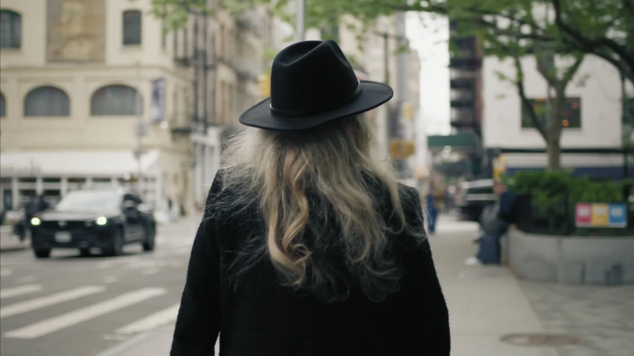 Faye (2024) movie still: medium — A woman walks away from the camera on a busy metropolitan sidewalk near cars. The blurred background shows city…