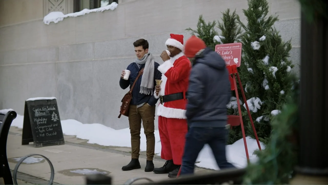 Ghosts of Christmas Past (2021) movie still: medium — A snowy outdoor sidewalk sits next to a concrete wall with Christmas-themed signage. A young…