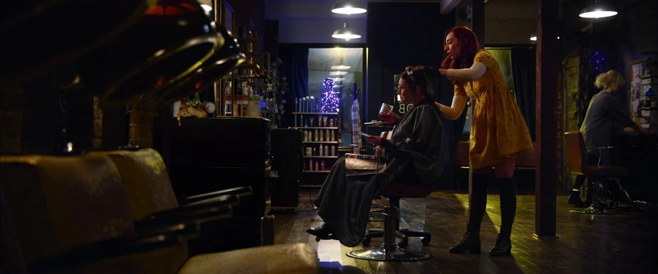 The Stylist (2020) movie still: medium — A dimly lit hair salon features brick walls and dark wooden floorboards. A stylist in a yellow dress…