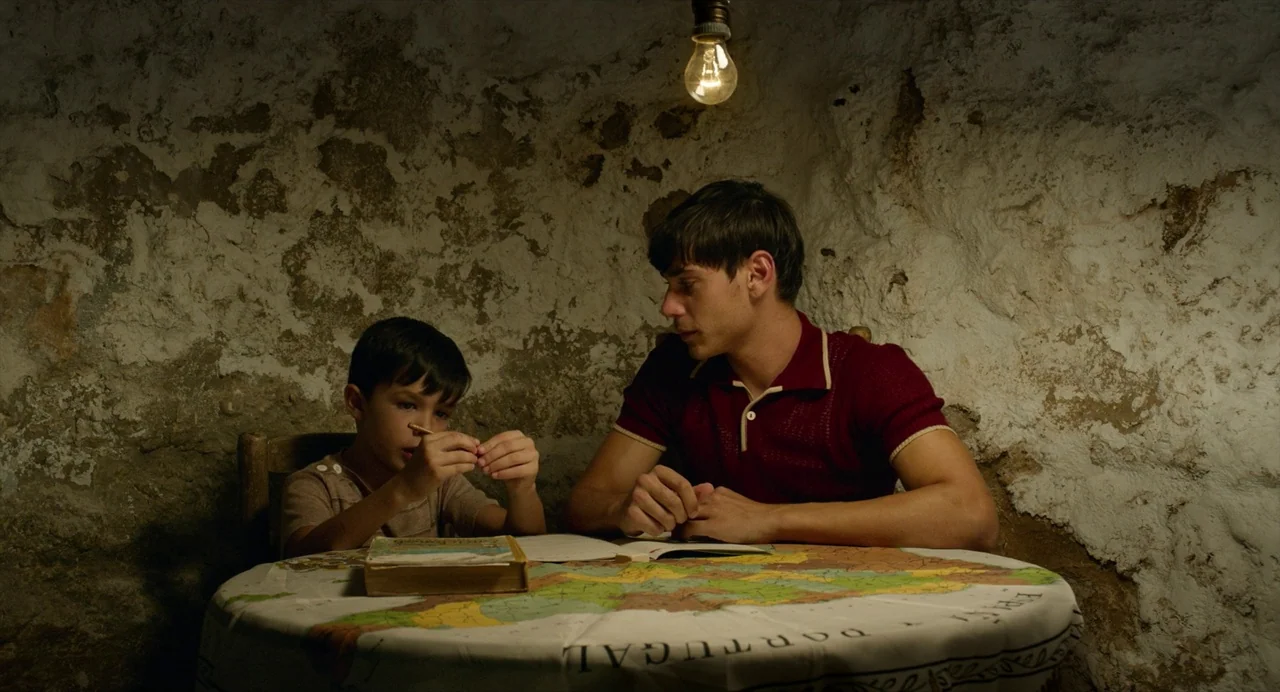 Pain and Glory (2019) movie still: medium — A rustic cellar with rough, uneven stone walls provides the backdrop for a table. Two males are seated…