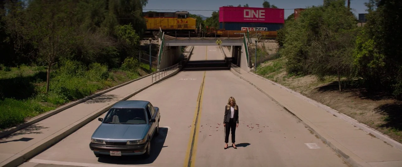 Promising Young Woman (2020) movie still: wide — A paved road passes beneath a railroad bridge where a train is crossing. A young woman stands alone…