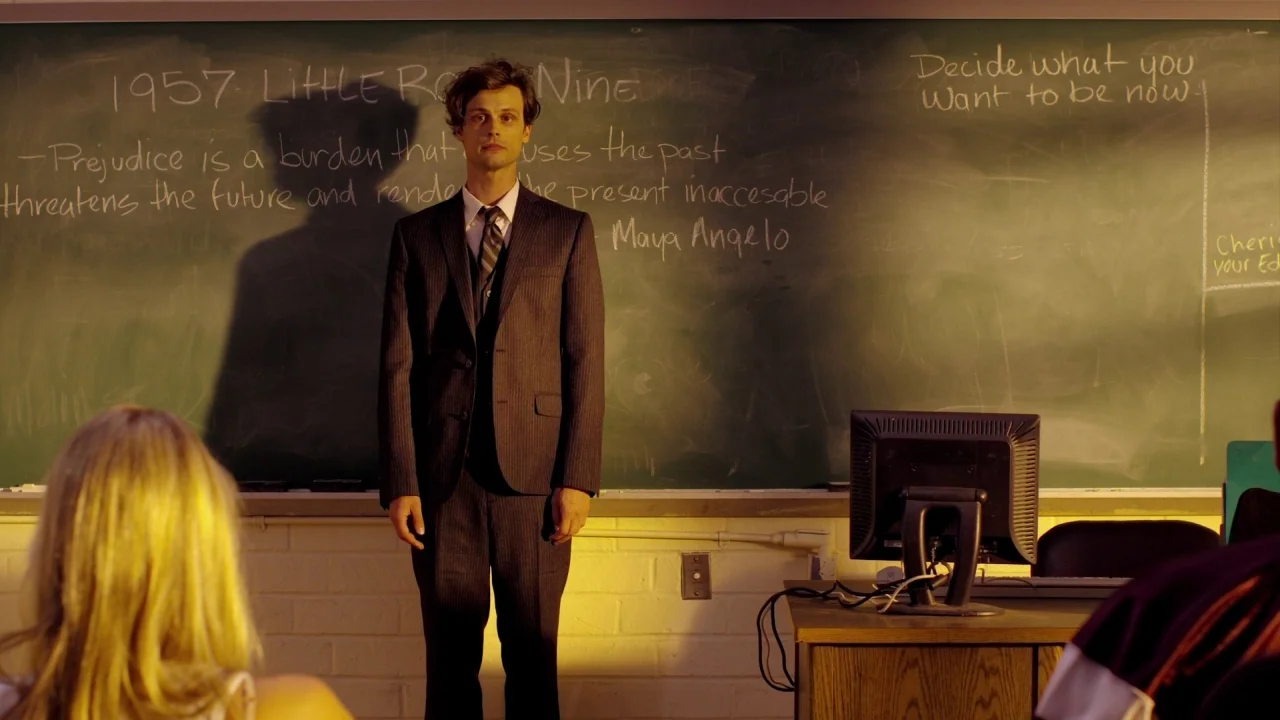 Suburban Gothic (2014) movie still: medium — A man in a suit stands before a chalkboard with handwritten educational text. A computer monitor sits…