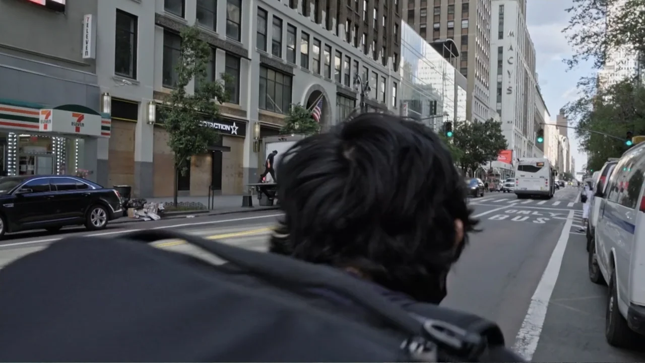 Covid Diaries NYC (2021) movie still: medium — A dense urban boulevard features tall historic buildings and storefronts under a daylight sky. A…