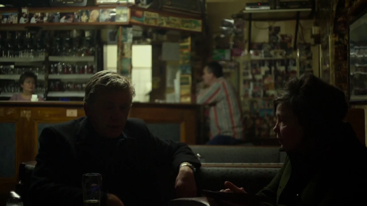 Museum Hours (2012) movie still: medium — An aging pub interior featuring wood-paneled walls and a cluttered bar background. An elderly man and…
