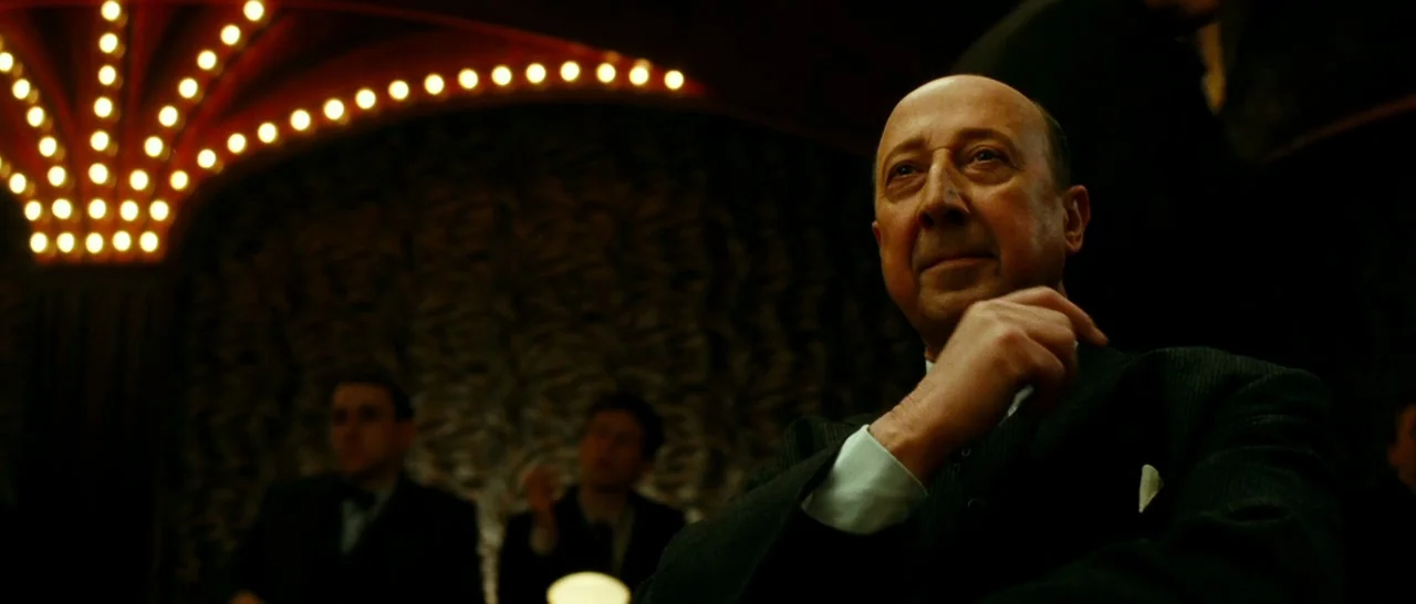 La Vie En Rose (2007) movie still: medium — A dimly lit club interior features rows of warm lights arched overhead. An elderly man in a suit sits in…
